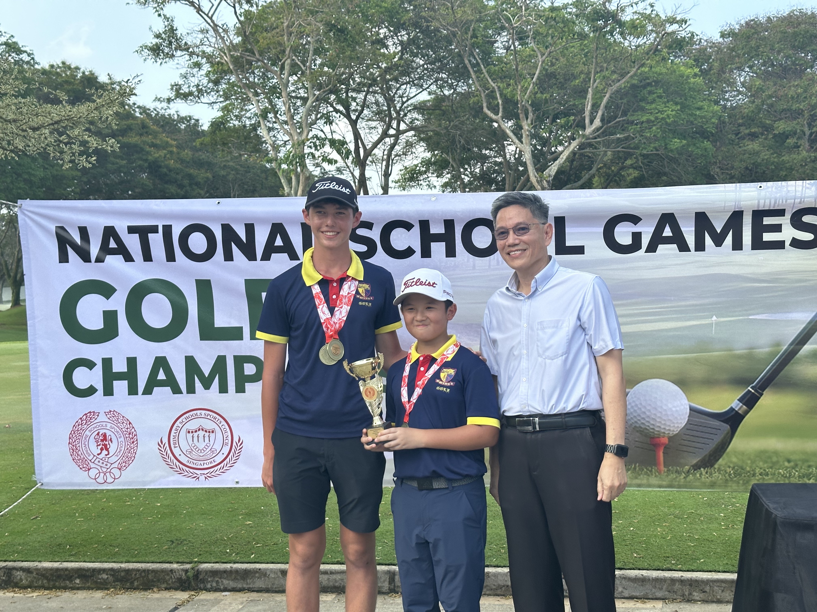 Two boys with medals and a trophy pose with an adult man before a National School Games Golf Championship banner.